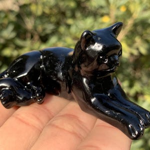 Könnte beinhalten: Glänzende schwarze Katzenfigur in Ruheposition. Die polierte Skulptur reflektiert das Licht und weist detaillierte Pfoten und Gesichtszüge auf. Die Katze liegt mit ausgestreckten Vorderpfoten. Dekoratives Objekt.