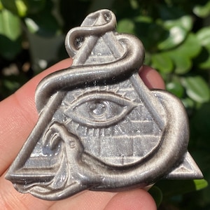 Könnte beinhalten: Ein silberfarbener Anhänger mit einem Auge in einem Dreieck, mit einer Schlange, die sich darum windet. Der Anhänger hat ein detailliertes Design und wird in einer Hand vor einem Hintergrund aus grünem Laub gehalten.