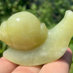 May include: A light green snail figurine carved from stone. The snail has a rounded shell and a smooth, curved body. The figurine is held in a hand against a blurred green background.