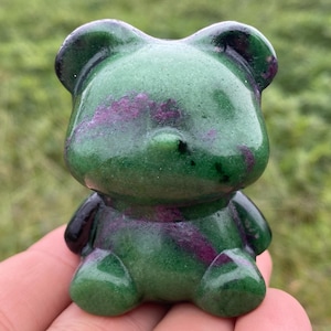 Pode incluir: Uma estatueta de urso verde com detalhes roxos e pretos. O urso tem um acabamento brilhante e é segurado em uma mão. A estatueta é um item decorativo, possivelmente um item de colecionador ou uma obra de arte única.
