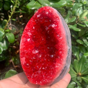 Könnte beinhalten: Eine oval geformte Geode mit einem leuchtend tiefroten kristallinen Inneren mit facettierten, funkelnden Texturen. Der äußere Rand des Steins zeigt ein glattes, hellgraues und durchscheinendes Mineralband, das das rote Zentrum einrahmt.