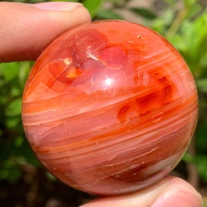 Peut inclure: Une sphère de cornaline polie et lisse, de couleur orange et rouge foncé. La sphère présente un motif tourbillonnant de bandes blanches et orange.