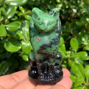 May include: A carved cat figurine made of green and black stone with a small pink inclusion. The cat is in a seated position with its tail curled up. The base of the figurine is black.
