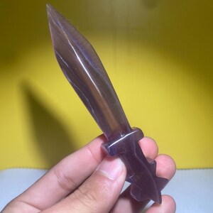 May include: A purple and black crystal sword with a detailed handle. The sword is made of polished stone and has a sharp point.