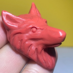 May include: A red carved wolf head pendant. The wolf's eyes are carved into the head and the fur is detailed.