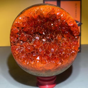 May include: A large, round, orange crystal geode with a hollow centre. The geode is on a red stand.