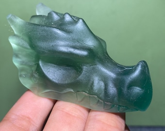 May include: A translucent, green dragon head sculpture. The dragon's features are detailed, with visible scales and a textured surface. The sculpture is held against a green background.