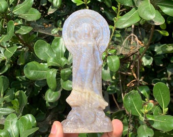 May include: A translucent, pale blue and tan carved stone figurine depicting a standing figure with a circular halo behind the head. The sculpture features intricate details of a draped garment and a base, showcasing a smooth, polished surface.