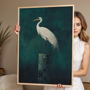 Könnte beinhalten: Gerahmter Kunstdruck mit einem weißen Reiher mit gelbem Schnabel und schwarzen Beinen, der auf einer verwitterten Steinsäule steht. Der Hintergrund ist ein dunkles Teal mit strukturiertem Aussehen, der Rahmen ist aus hellem Holz.