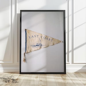May include: A triangular off-white pennant flag framed in black, featuring a blue illustration of a sailboat and the text EAST COAST printed in blue. The fabric has a weathered texture with a small rope tassel attached to the left side.
