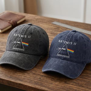 Può includere: Due cappellini da baseball, uno grigio antracite e uno blu navy, sono esposti su una superficie di legno. Ogni cappellino presenta la scritta "ARTEMIS II" sopra una grafica di un prisma e arcobaleno, con "DARK SIDE OF THE MOON" sotto. I cappellini hanno un aspetto vintage.
