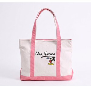 May include: A white canvas tote bag with pink trim and handles. The front features a graphic of Mickey Mouse drawing a black line that leads to the text "Mrs. Watson" in a stylized black font. The bag has a rectangular shape and pink base.