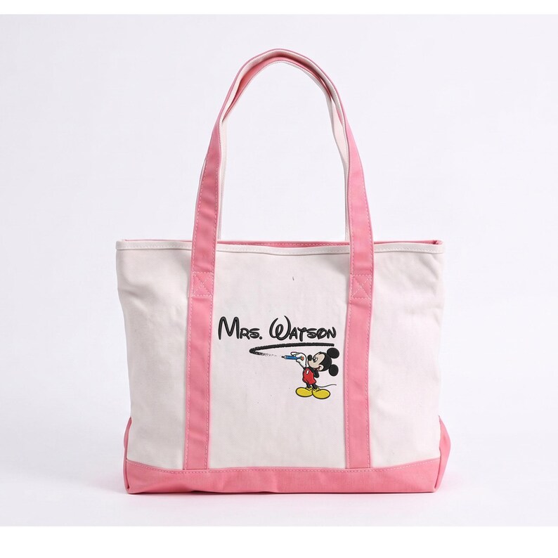May include: A white canvas tote bag with pink trim and handles. The front features a graphic of Mickey Mouse drawing a black line that leads to the text "Mrs. Watson" in a stylized black font. The bag has a rectangular shape and pink base.