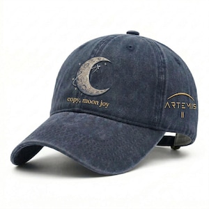 Puede incluir: Gorra de béisbol azul marino con visera curva. La gorra presenta un gráfico de media luna con el texto "copy, moon joy" y "ARTEMIS II" en letras doradas. La gorra tiene una correa ajustable.