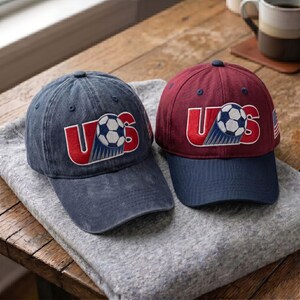 K&ouml;nnte beinhalten: Zwei Baseballkappen mit dem Schriftzug US, bei dem ein Fu&szlig;ball den Buchstaben S ersetzt. Die linke Kappe ist in verwaschenem Dunkelblau gehalten, die rechte hat eine kastanienbraune Krone und einen dunkelblauen Schirm. Beide haben eine kleine Flagge.