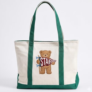 K&ouml;nnte beinhalten: Cremefarbene Canvas-Tragetasche mit gr&uuml;nen Akzenten und Griffen. Sie zeigt einen gestickten braunen Teddyb&auml;ren, der ein Banner mit der Aufschrift "STANFORD" h&auml;lt. Die Tasche hat eine rechteckige Form.