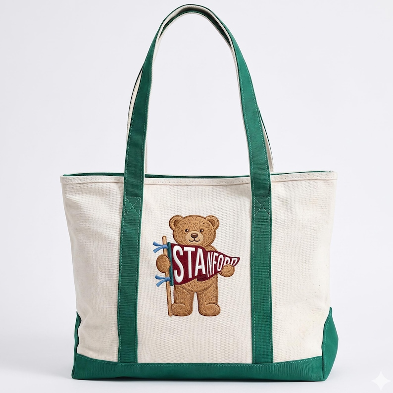 K&ouml;nnte beinhalten: Cremefarbene Canvas-Tragetasche mit gr&uuml;nen Akzenten und Griffen. Sie zeigt einen gestickten braunen Teddyb&auml;ren, der ein Banner mit der Aufschrift "STANFORD" h&auml;lt. Die Tasche hat eine rechteckige Form.