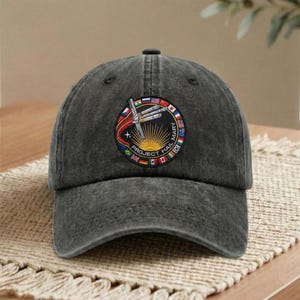 May include: A dark gray baseball cap with a circular patch featuring a spacecraft, flags, and the text "PROJECT HAIL MARY." The patch is surrounded by a ring of flags. The cap has a curved brim and a button on top.