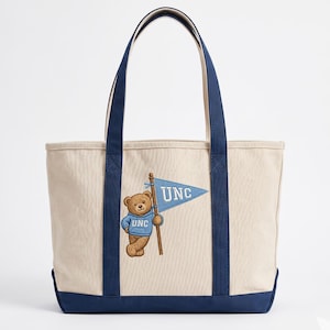 May include: A beige tote bag with navy blue accents, featuring a cartoon bear holding a flag that reads "UNC." The bear wears a light blue hoodie with "UNC" embroidered on it. The bag has navy blue handles and a navy blue base.