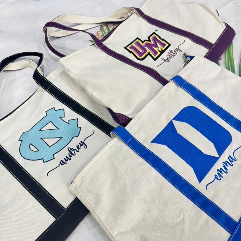 K&ouml;nnte beinhalten: Off-white Canvas-Tragetaschen mit College-Logos in Blau, Lila und Schwarz. Jede Tasche hat eine kontrastierende Bord&uuml;re und Griffe. Die Namen "Audrey", "Brantley" und "Emma" sind in Schreibschrift auf den Taschen geschrieben.