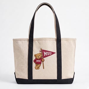 May include: Cream-colored tote bag with black handles and base. The bag features an embroidered design of a teddy bear wearing a maroon sweater and holding a maroon pennant with "MSU" in white letters. The bag is made of canvas.