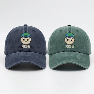 May include: Two baseball caps, one navy blue and one green, are displayed. Each cap features an embroidered design of a smiling face with a globe on its head, and the word "RISE" below. The caps are made of a soft, textured fabric.