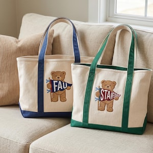 K&ouml;nnte beinhalten: Zwei cremefarbene Canvas-Tragetaschen mit gestickten Teddyb&auml;ren, die Wimpel halten. Eine Tasche hat marineblaue Griffe und Verzierungen, die andere gr&uuml;ne. Die Wimpel tragen die Aufschrift "FAU" und "Stanford".