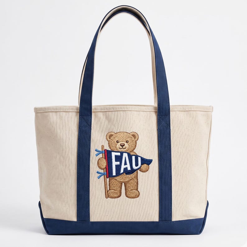 K&ouml;nnte beinhalten: Beige Canvas-Tragetasche mit marineblauen Akzenten. Mit einem gestickten braunen Teddyb&auml;ren, der eine Flagge mit den wei&szlig;en Buchstaben "FAU" h&auml;lt. Die Tasche hat marineblaue Griffe und einen marineblauen Boden.