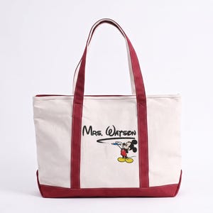 May include: A cream-colored canvas tote bag with dark red trim and handles. The front features the black text Mrs. Watson written in a cursive font, with a graphic of Mickey Mouse holding a blue pen and drawing a black line underneath.