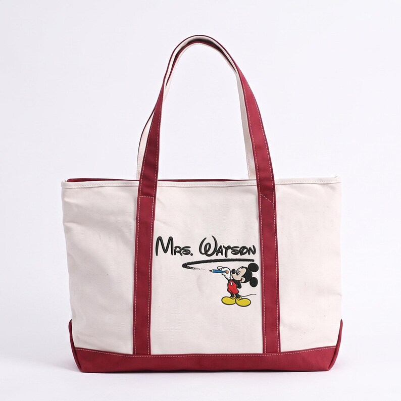May include: A cream-colored canvas tote bag with dark red trim and handles. The front features the black text Mrs. Watson written in a cursive font, with a graphic of Mickey Mouse holding a blue pen and drawing a black line underneath.