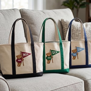 May include: Three canvas tote bags with different colored accents and handles. Each bag features a teddy bear holding a pennant with a university's initials: MSU, UCB, and UNC. The bags are beige with black, green, and navy blue accents.