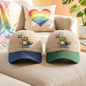 Può includere: Due cappellini da baseball con corona beige e una grafica di rana supereroe che tiene una bandiera arcobaleno. Un cappellino ha la visiera blu navy, l'altro verde. Un cuscino a forma di cuore arcobaleno e una bandiera arcobaleno sullo sfondo.