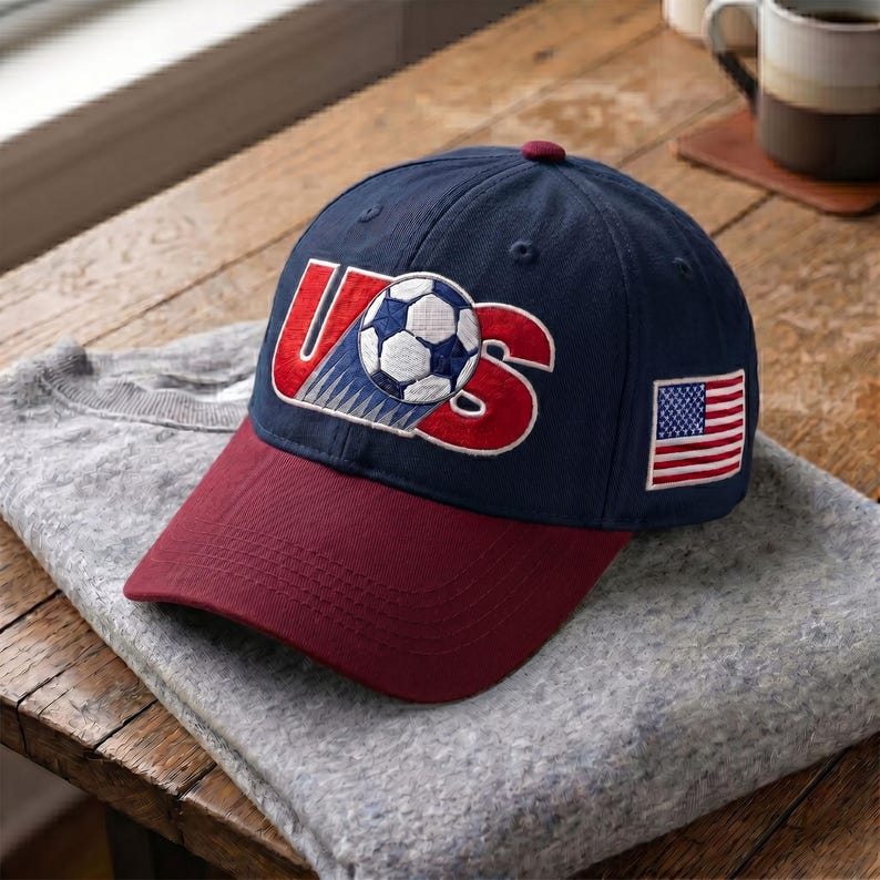 K&ouml;nnte beinhalten: Eine marineblaue Baseballkappe mit bordeauxrotem Schirm. Auf der Vorderseite befindet sich ein gesticktes rotes 'US'-Logo, bei dem ein Fu&szlig;ball den Buchstaben 'S' ersetzt. Ein kleiner amerikanischer Flaggenaufn&auml;her ist seitlich aufgestickt.