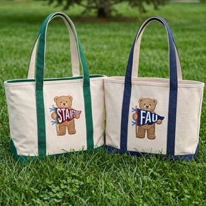 K&ouml;nnte beinhalten: Zwei Canvas-Tragetaschen mit gestickten Teddyb&auml;ren, die Wimpel halten. Eine Tasche hat gr&uuml;ne Griffe und Verzierungen mit "STANFORD" auf dem Wimpel. Die andere hat marineblaue Griffe und Verzierungen mit "FAU" auf dem Wimpel.