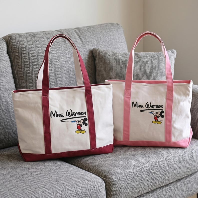 May include: Two off-white canvas tote bags with dual handles. The left bag features dark red trim and the text "Mrs. Watson" with a graphic of Mickey Mouse drawing the line. The right bag has light pink trim and identical text and graphic.
