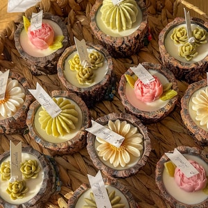 Pode incluir: Uma coleção de velas decorativas em suportes de madeira rústicos, cada uma encimada por um design floral diferente. As velas apresentam uma variedade de cores, incluindo rosa, amarelo e verde, e estão dispostas numa cesta de vime. Cada vela tem uma pequena etiqueta com "Lumila".