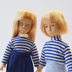 May include: Two vintage dolls with blonde hair and blue eyes. They wear matching blue and white striped tops. One doll is wearing a navy blue skirt, and the other is wearing light blue pants. The dolls are standing side by side.