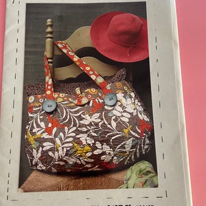 May include: A quilted travel bag with a floral pattern, measuring 22 inches by 17 inches, is displayed with a red hat. The bag has two large wooden buttons and a patterned strap. The text "ON THE ROAD AGAIN - PART I" is visible.