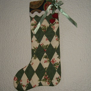 May include: A quilted Christmas stocking with a green and cream diamond pattern, adorned with holly and red berries. The top features a paisley fabric and a green satin ribbon with two red heart-shaped ornaments.