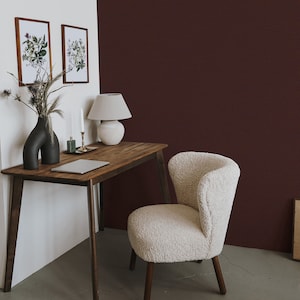 May include: A wooden desk with a white fluffy chair, a laptop, a white lamp, and a vase with a plant. The desk is against a white wall and a dark red wall.