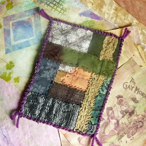May include: A handmade patchwork textile with a variety of fabrics in shades of green, brown, and orange, stitched together. The edges are finished with a purple yarn, and the corners have small yarn loops. The background includes vintage paper.