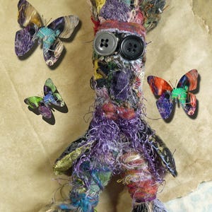 May include: A handmade textile doll with button eyes and a multi-coloured, textured body. The doll is surrounded by three decorative butterflies with colourful patterns. The background is a textured, tan surface.