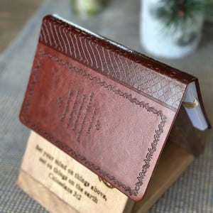 May include: A brown leather-bound journal with a decorative embossed design and the quote "Set your mind on things above, not on things on the earth. Colossians 3:2" on the front cover.
