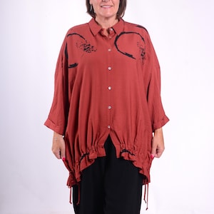 May include: A rust-colored button-down shirt with black abstract designs and a gathered hemline. The shirt is paired with black pants and silver sandals. The model is standing in front of a white background.