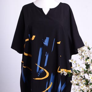 Pode incluir: Top preto estilo túnica com decote em V e mangas curtas. O top apresenta um design abstrato arrojado em pinceladas azuis e amarelas. A peça de roupa é exibida num manequim sobre um fundo branco, com flores brancas no canto.