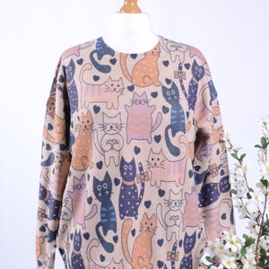 Könnte beinhalten: Beiger Rundhals-Pullover mit einem sich wiederholenden Muster aus Cartoon-Katzen in Orange, Blau und Lila. Die Katzen haben verschiedene Posen, mit kleinen schwarzen Herzen dazwischen. Der Pullover wird auf einer Schaufensterpuppe vor weißem Hintergrund präsentiert.