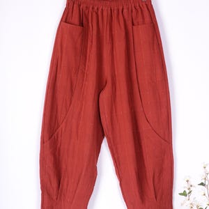 May include: Rust-colored, loose-fitting pants with large front pockets, hanging on a wooden hanger. The fabric has a subtle grid pattern. The pants have a relaxed, comfortable style, suitable for casual wear.