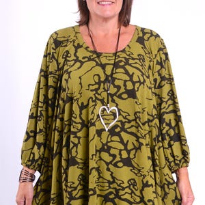 Peut inclure: Haut tunique vert olive avec un motif abstrait noir. Le haut a des manches longues et une encolure arrondie. Un pendentif en forme de cœur argenté est suspendu à un collier en cordon noir. Un bracelet argenté est porté.