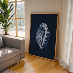 May include: A framed print featuring a white seashell design on a navy blue background. The artwork is displayed in a room with a gray sofa, a potted plant, and a window with natural light. The frame is made of light-colored wood.