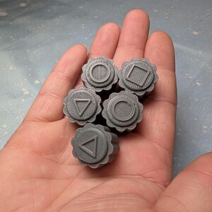 May include: Five gray plastic buttons with different symbols: a circle, a square, a triangle, and two upside-down triangles.